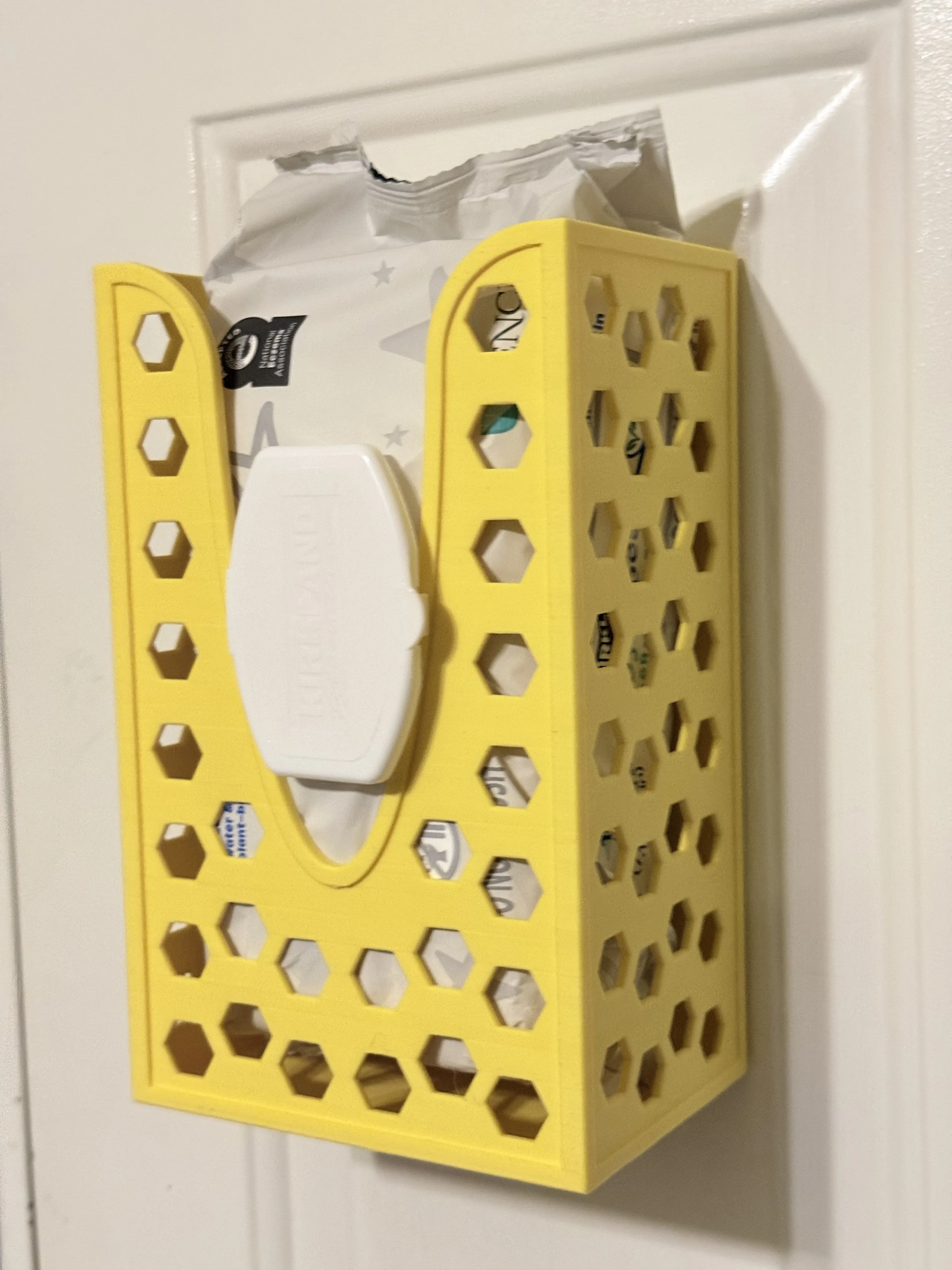 A yellow dispenser mounted to a white metal door, holding a heavy pack of Kirkland baby wipes. This photo demonstrates the dispenser's high-friction mounting system supporting significant weight without sliding.