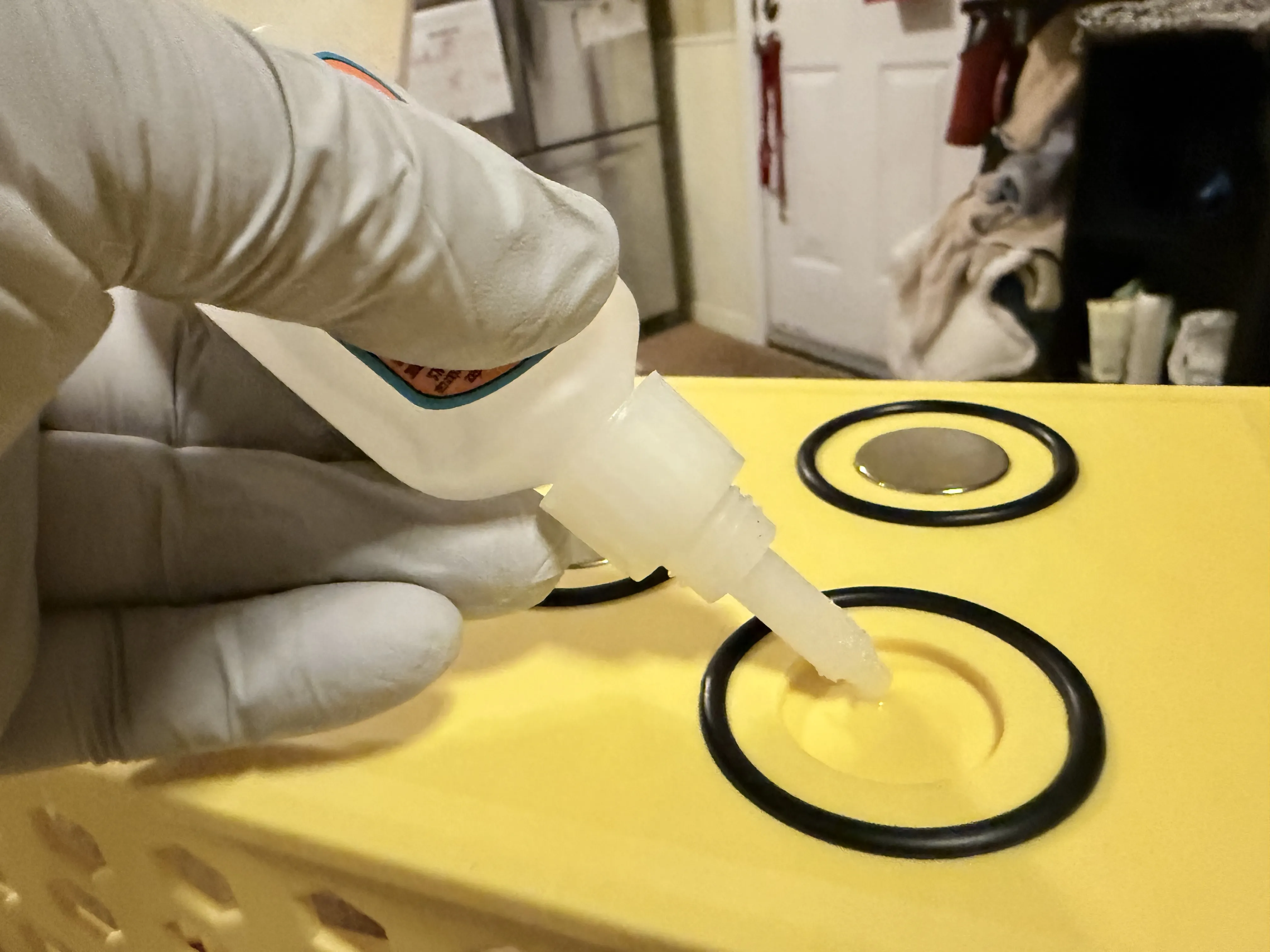 A gloved hand applying liquid adhesive from a precision tip bottle into the center of a circular magnet recess. The recess is already encircled by a black rubber O-ring, and one magnet is visible already glued into an adjacent slot.