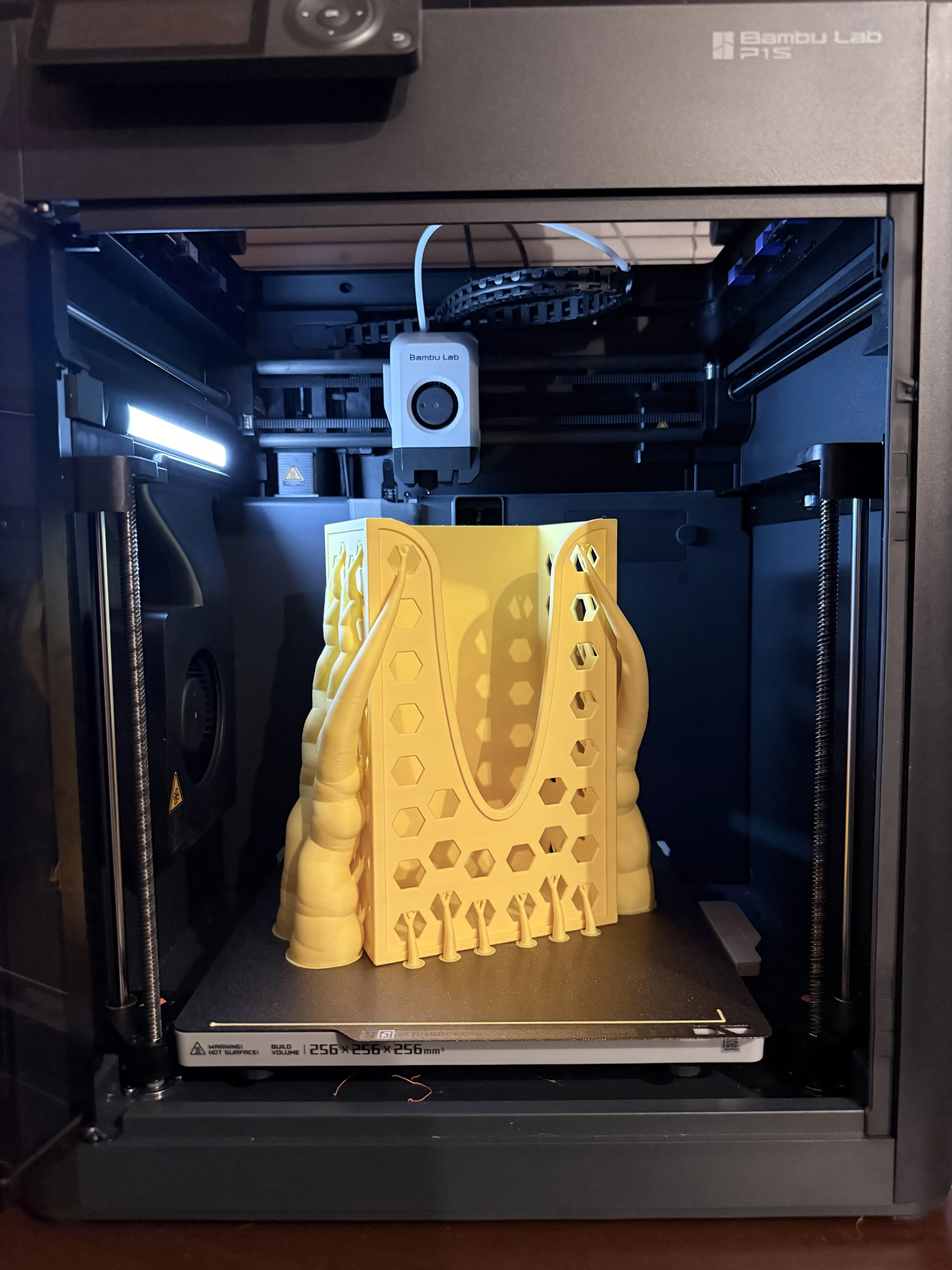 A photograph of the completed 3D printed glove dispenser in yellow filament, sitting on the build plate of a Bambu Lab P1S. The print is shown with the organic tree supports still attached, which branch out from the base to support the front ergonomic cutout and side hexagonal patterns.