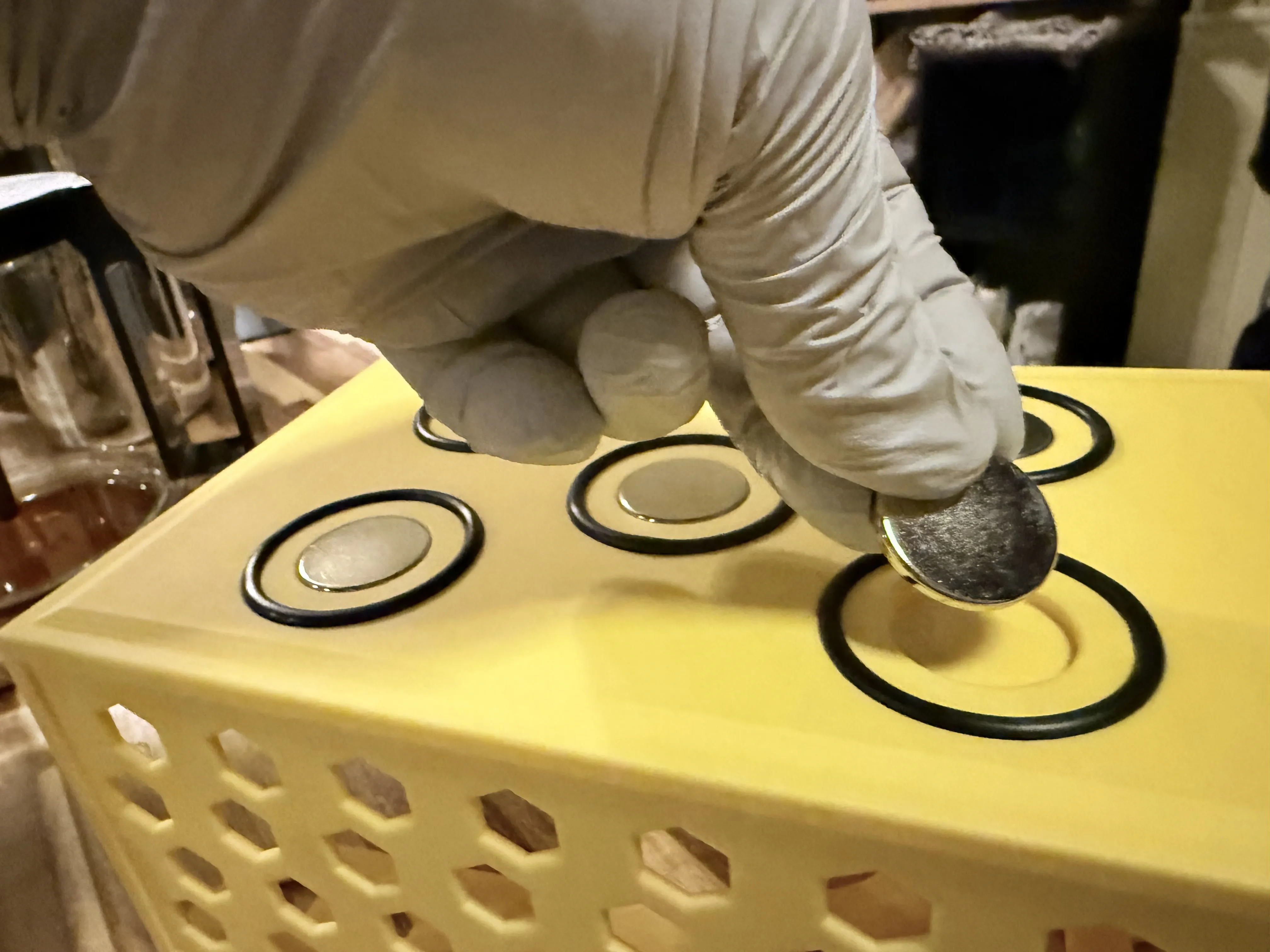 A gloved hand carefully placing a silver 25mm neodymium magnet into a circular recess on the back of the yellow dispenser. The recess is surrounded by a black O-ring, and three other magnets are already visibly installed in the nearby slots.
